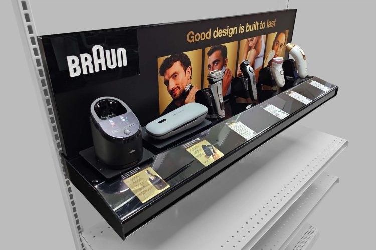 Black gloss–powder-coated steel retail display prototype for Braun grooming devices. Five products (shavers, trimmers, laser hair removal handset) are mounted on clear PETG brackets. Below each device, a clear acrylic channel holds a product information card. The magnetic back panel features raised white acrylic “Braun” letters on the left and the gold tagline “Good design is built to last.”