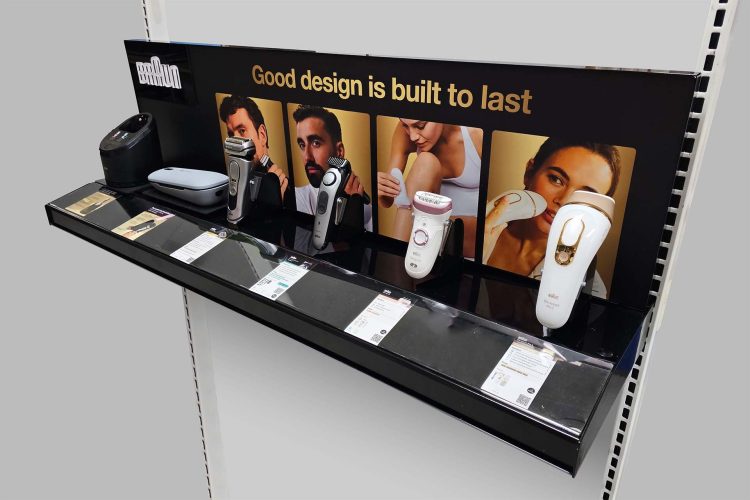 Black gloss–powder-coated steel retail display prototype for Braun grooming devices. Five products (shavers, trimmers, laser hair removal handset) are mounted on clear PETG brackets. Below each device, a clear acrylic channel holds a product information card. The magnetic back panel features raised white acrylic “Braun” letters on the left and the gold tagline “Good design is built to last.”