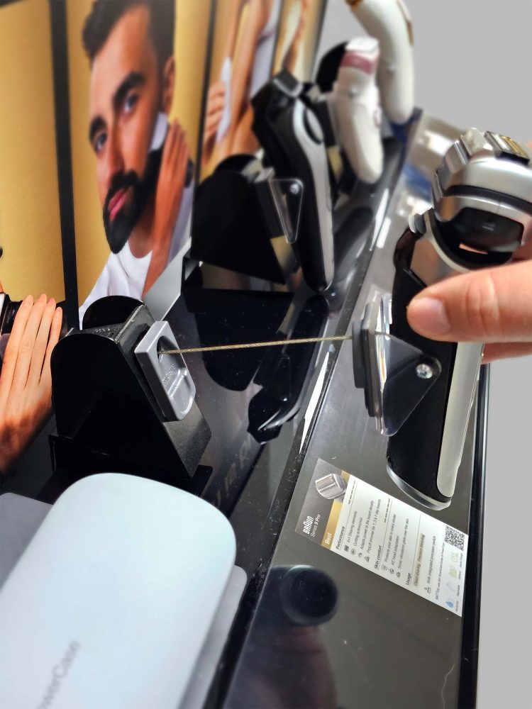 Retail display featuring Braun electric shavers mounted on clear stands. A hand is interacting with one of the devices. Behind the shavers, lifestyle images show people using the products. An informational card highlights features of a specific shaver, including branding and product details.