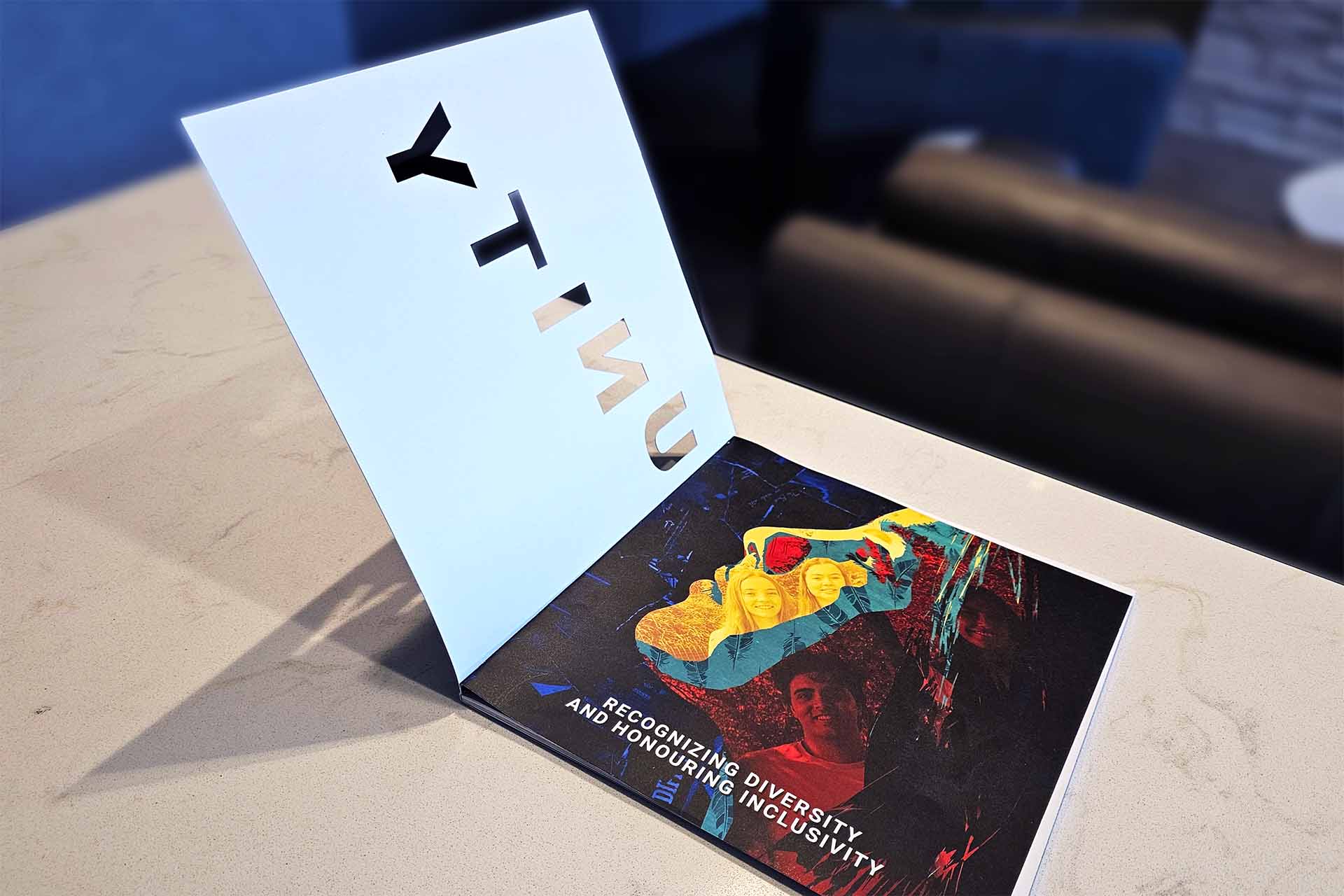 Open booklet showing cut-out letters spelling “UNITY” and a collage of diverse faces with text about inclusivity