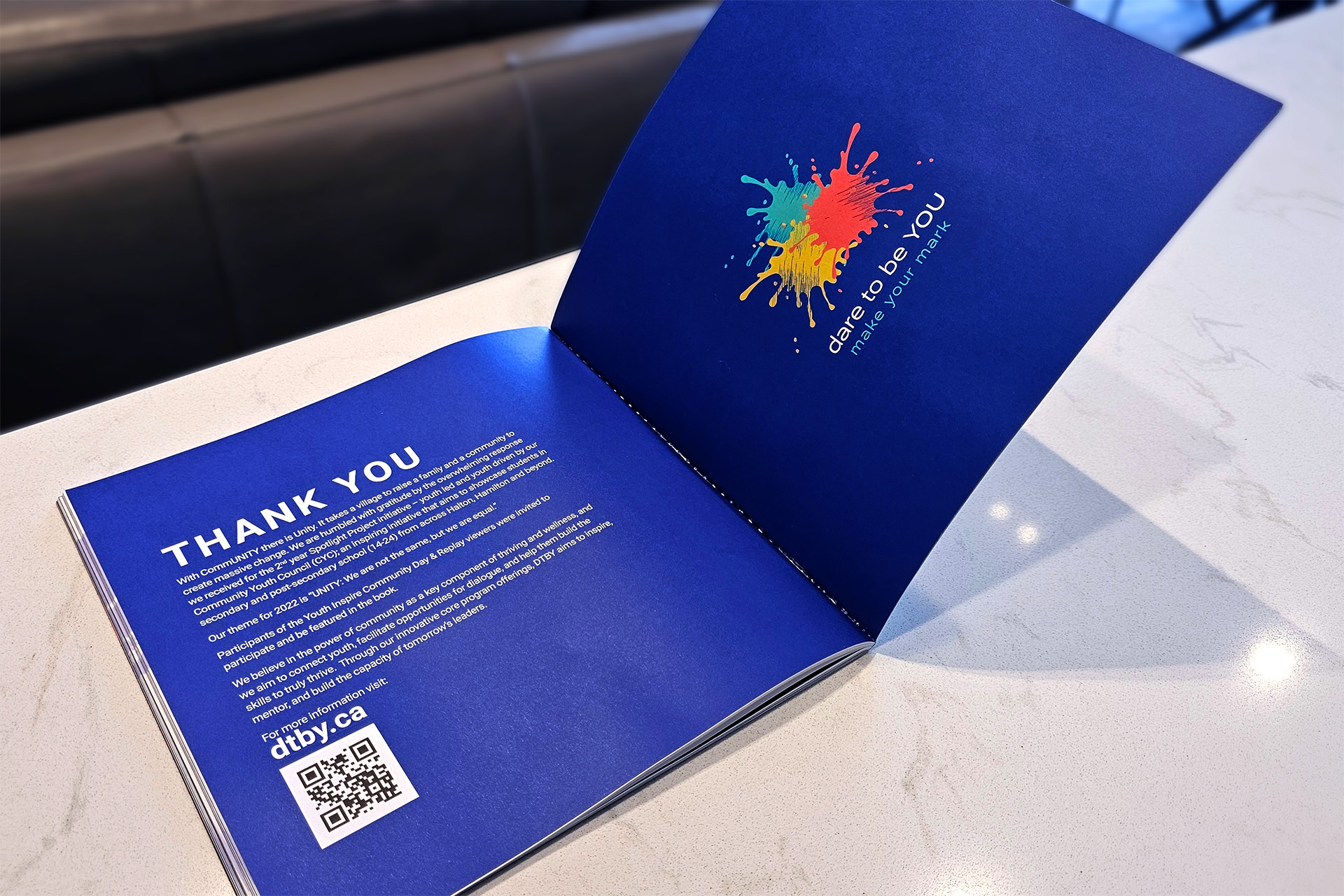 Open booklet showing a thank-you message on one page and colorful paint splatter with text “dare to be YOU make your mark” on the other.