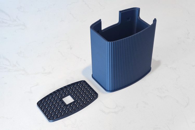 The blue ridged shroud with its open end facing up, next to a black gyroid-infill dust screen with a square cutout, on a white marble counter.