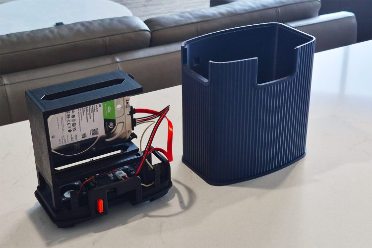 Open black chassis with an 8 TB Seagate BarraCuda drive in its caddy, red and yellow cables and an orange power button, beside the empty blue ridged shroud on a white counter.